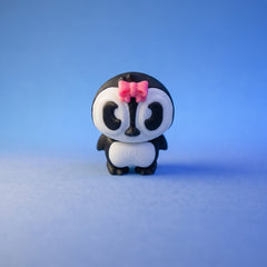 Dottie penguin figurine standing alone with a pink bow on her head against a blue background.