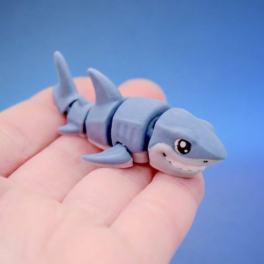 Grey 3D-printed great white shark fidget toy with a cheeky grin and white belly, curled in a hand.